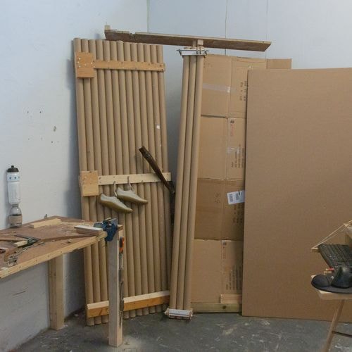 Een rommelige werkplaats met een werkbank vol gereedschap, stapels kartonnen buizen met hangende schoenen en kartonnen dozen.