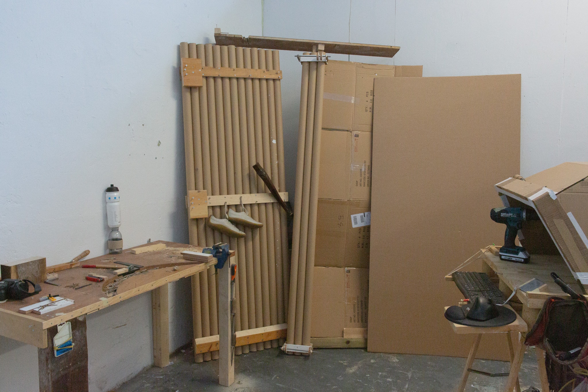 Een rommelige werkplaats met een werkbank vol gereedschap, stapels kartonnen buizen met hangende schoenen en kartonnen dozen.