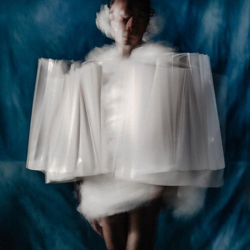 Een bewegende figuur in een volumineus wit, wolkachtig en geplooid kledingstuk staat voor een donkerblauwe achtergrond.