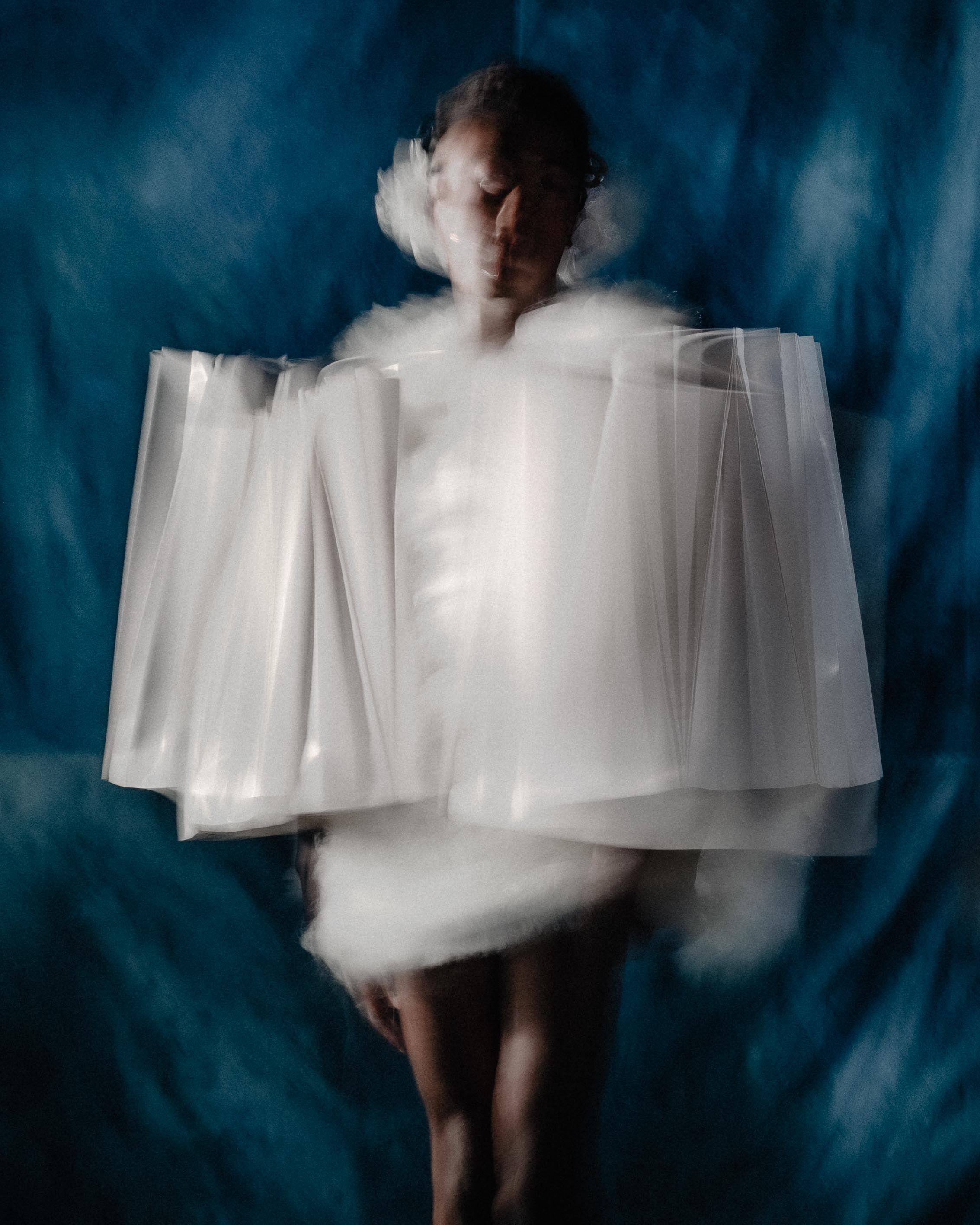 Een bewegende figuur in een volumineus wit, wolkachtig en geplooid kledingstuk staat voor een donkerblauwe achtergrond.