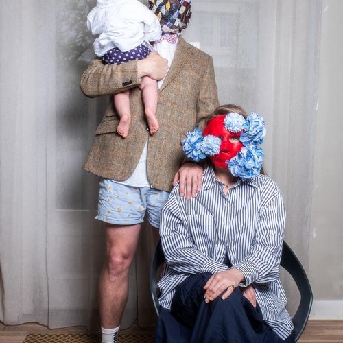 Familieportret: vader met stropdassenmasker houdt baby vast, moeder met rood bloemenmasker zit in stoel.