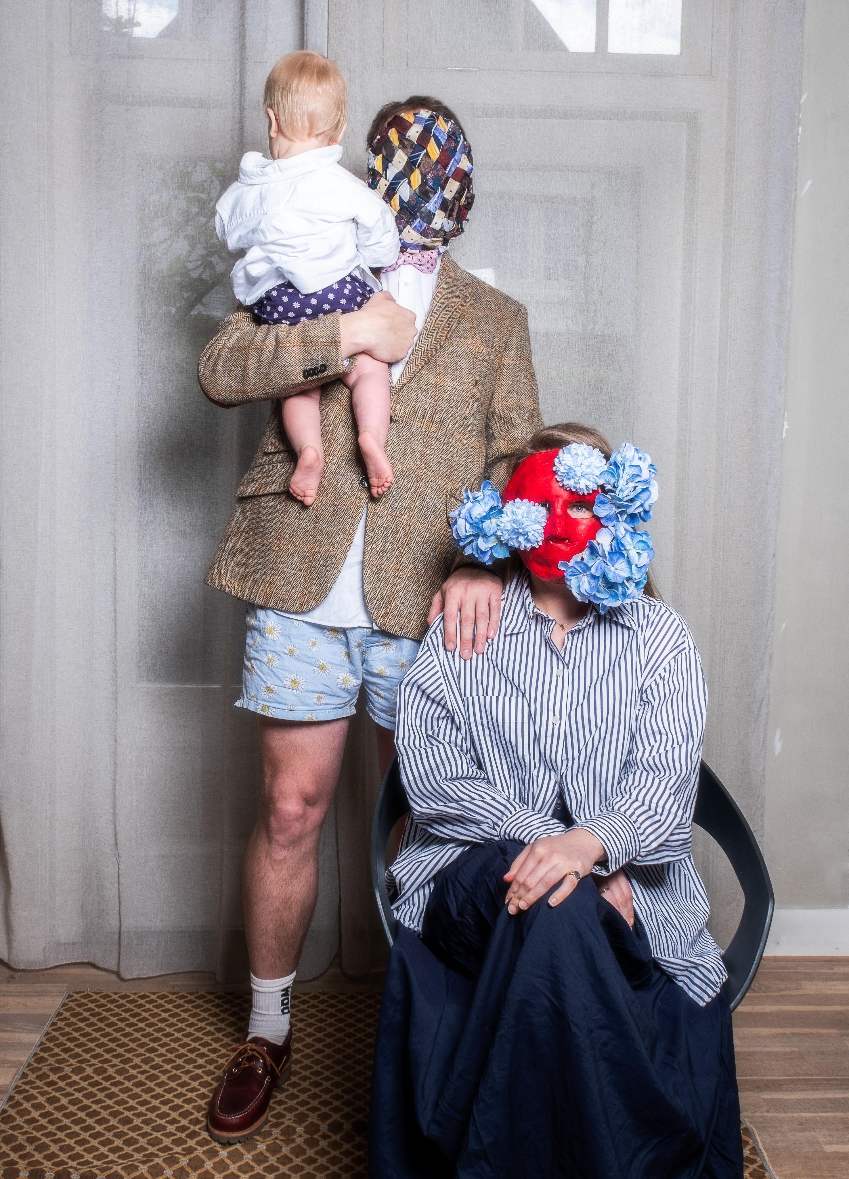 Familieportret: vader met stropdassenmasker houdt baby vast, moeder met rood bloemenmasker zit in stoel.