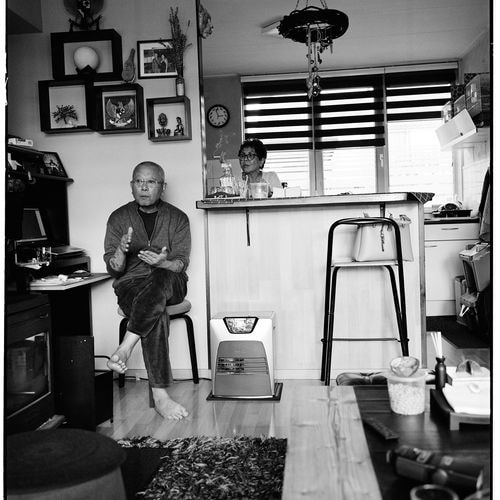 A bald man with glasses and tattoos gestures from a stool, barefoot, to a woman behind the counter in a busy room.