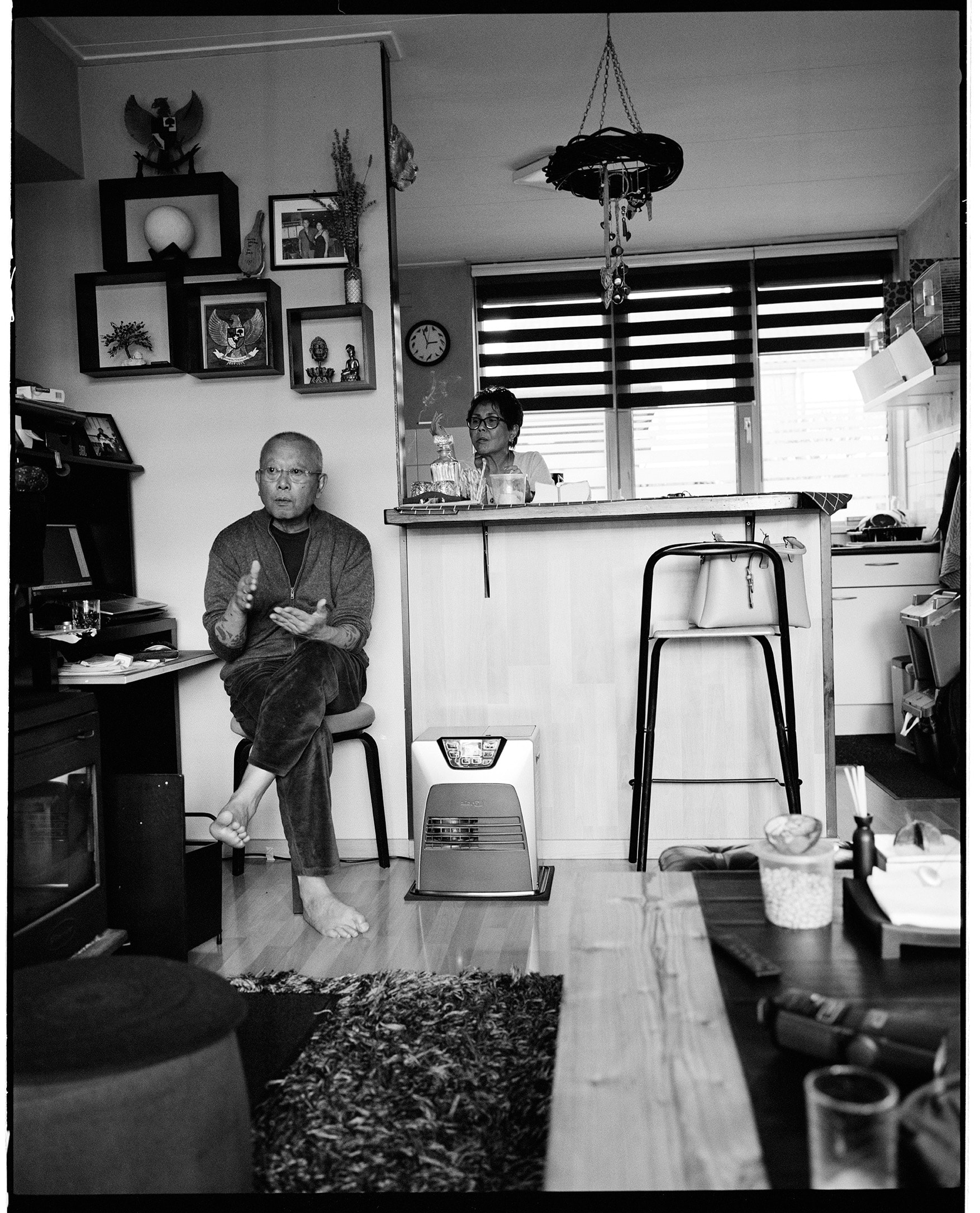 Een kale man met een bril en tatoeages gebaart vanaf een kruk, op blote voeten, naar een vrouw achter de toonbank in een drukke ruimte.