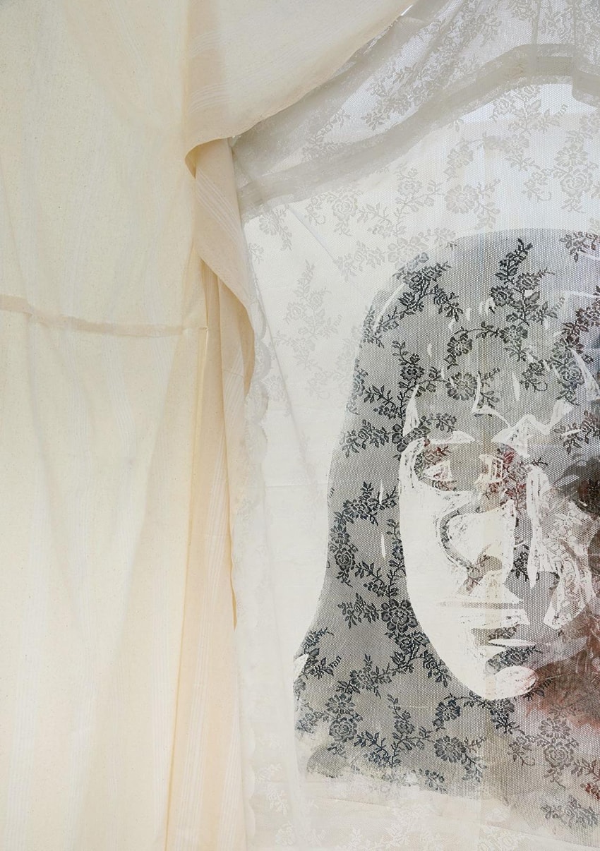 A woman's face is visible through a sheer white lace veil with dark floral motifs, framed by cream-colored curtains.