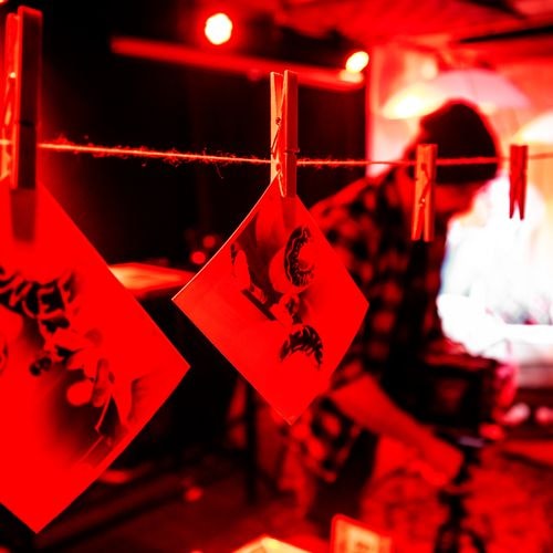 Twee roodgetinte foto's met abstracte, donkere vormen hangen aan een touwtje, vastgebonden met wasknijpers. Een wazig persoon staat in een rood verlichte kamer.