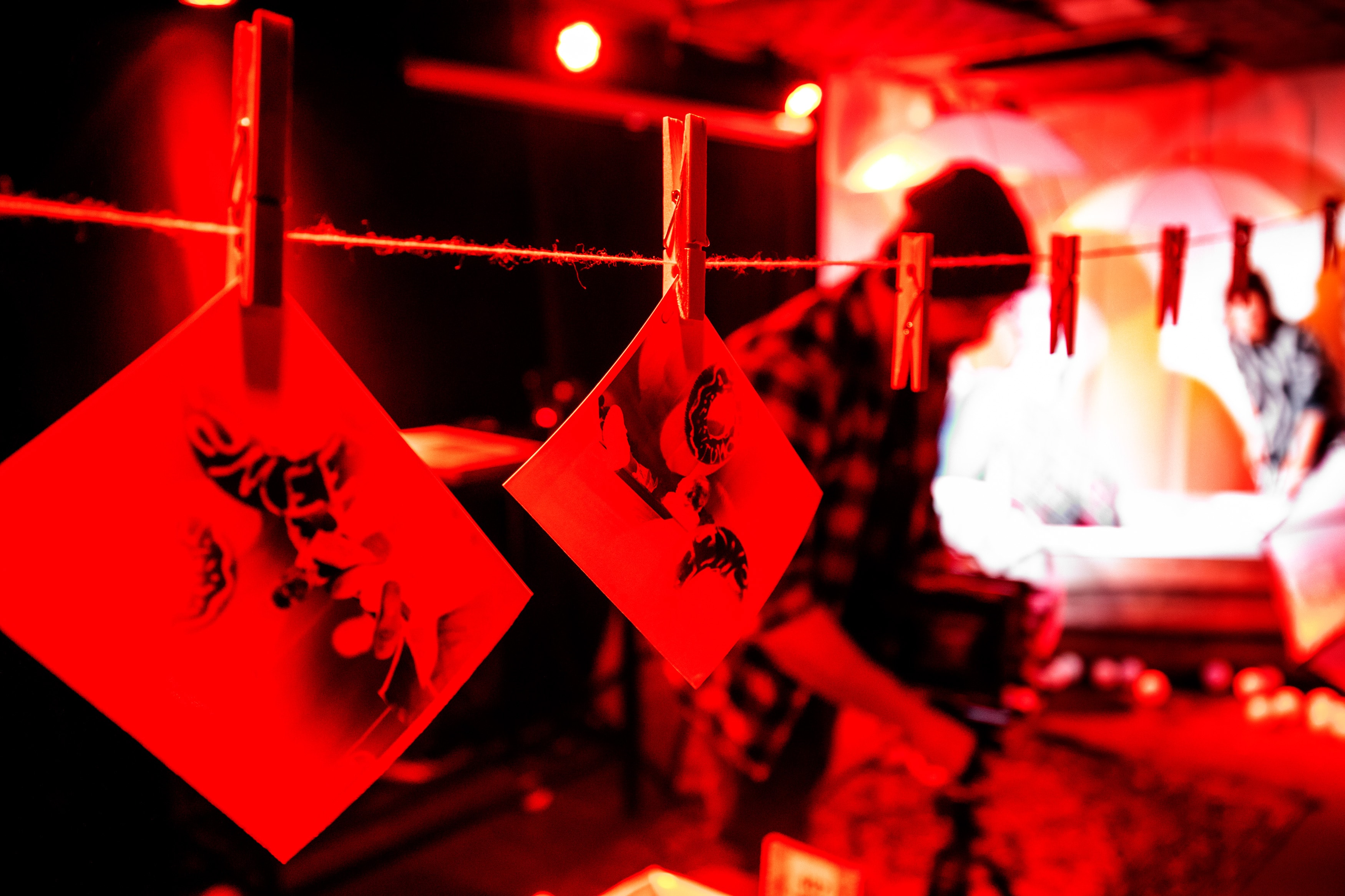 Twee roodgetinte foto's met abstracte, donkere vormen hangen aan een touwtje, vastgebonden met wasknijpers. Een wazig persoon staat in een rood verlichte kamer.
