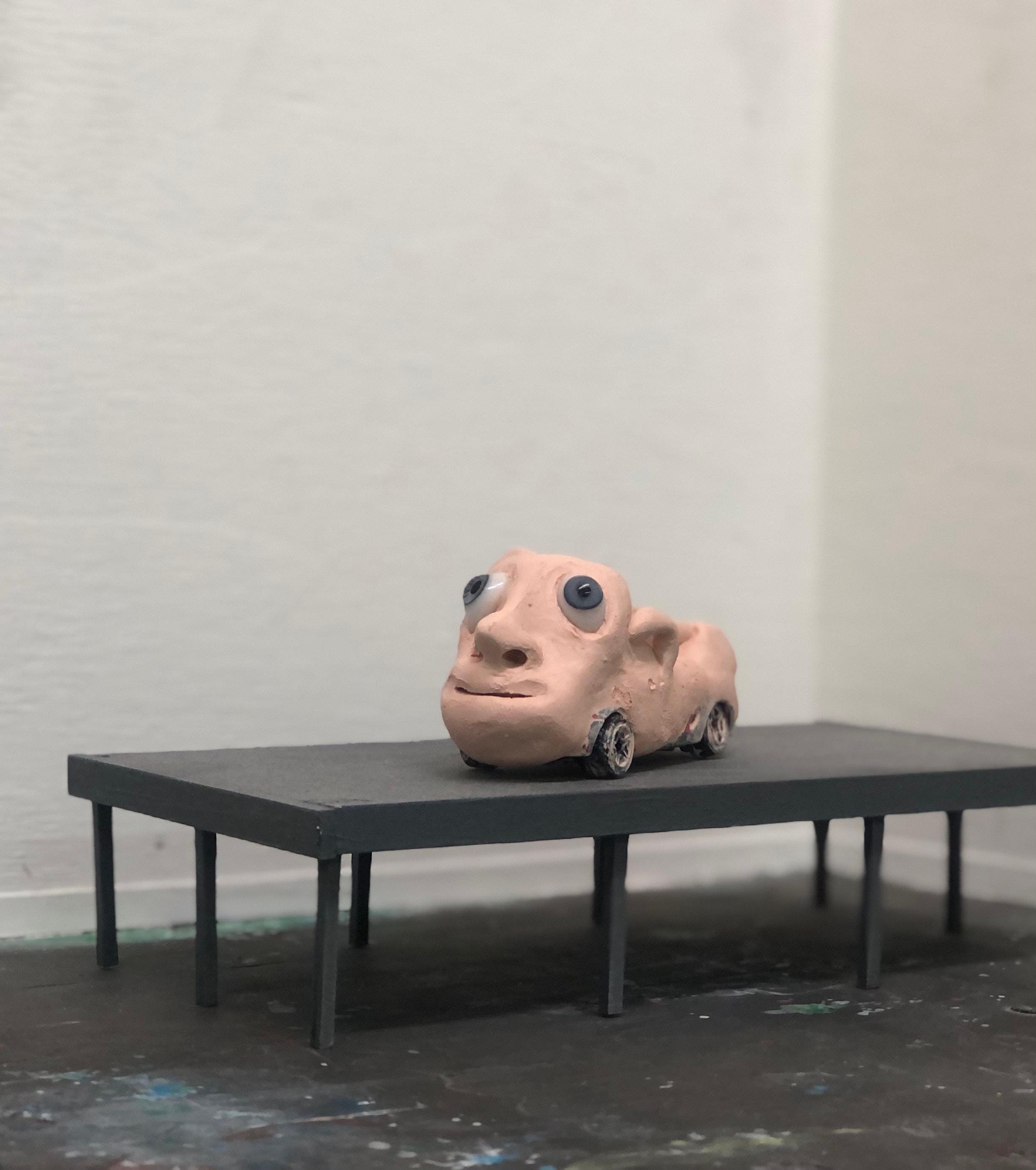 Een vreemd, vleeskleurig kleifiguur: een gezicht met grote ogen, op een pottenbakkerswiel, op een donkergrijze tafel voor een witte muur.