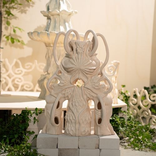 Een sierlijke, lichtgekleurde keramische sculptuur met abstracte gravures staat op bakstenen, tegen een wazige, gelaagde fontein en groene planten op de achtergrond.