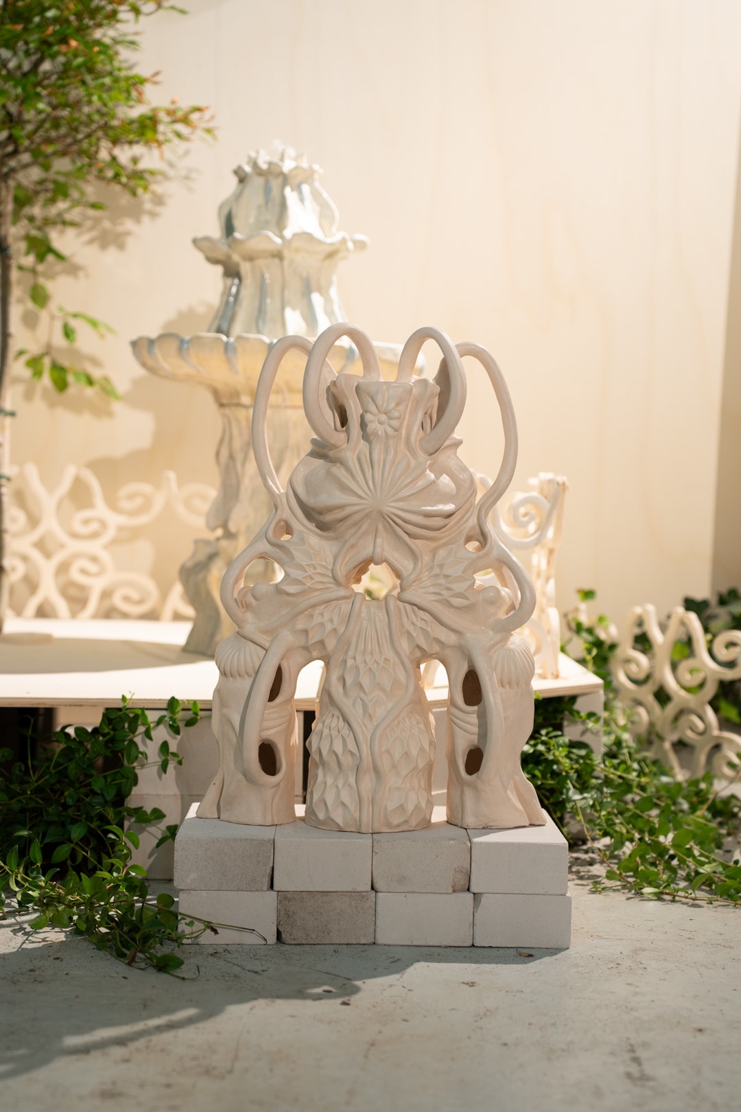 Een sierlijke, lichtgekleurde keramische sculptuur met abstracte gravures staat op bakstenen, tegen een wazige, gelaagde fontein en groene planten op de achtergrond.