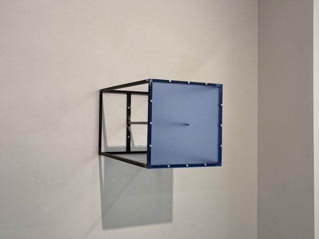 Blauw doorschijnend paneel met donkere kegel in zwart kubusframe, aan een witte muur bevestigd.