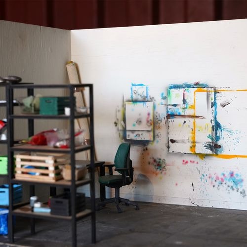 Miniatuurkunstenaarsatelier met een boekenkast, bureaustoel en witgekalkte muren bedekt met kunstwerken.