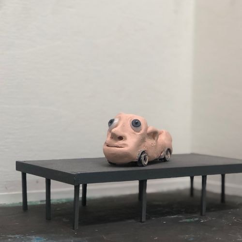 Een vreemd, vleeskleurig kleifiguur: een gezicht met grote ogen, op een pottenbakkerswiel, op een donkergrijze tafel voor een witte muur.